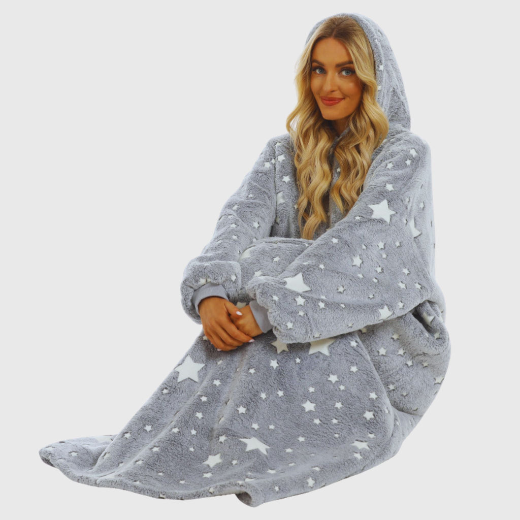 Woman sitting cross-legged in a grey glow-in-the-dark star print hoodie blanket from Hoodie Haven, smiling in a cozy, relaxed pose.